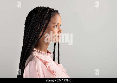 Vista laterale della giovane ragazza afro-americana in blusa rosa vintage con lunghi capelli intrecciati che guardano lontano da sfondo grigio Foto Stock