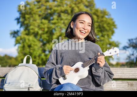 Bella giovane donna seduta con zaino su panchina in parco soleggiato, suona la chitarra ukulele e canta lungo Foto Stock