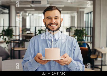 Uomo eccitato che celebra il compleanno in ufficio, cercando felice, tenendo la torta con la candela, facendo un desiderio. Foto Stock