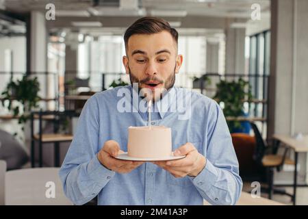 Uomo eccitato che celebra il compleanno in ufficio, cercando felice, tenendo la torta con la candela, facendo un desiderio. Foto Stock