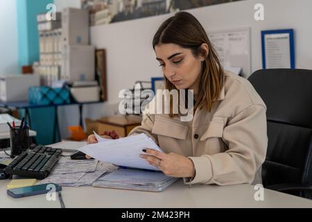 beauty giovane donna in tuta scrivere su un blocco note e telefonare mentre si siede in un ufficio Foto Stock