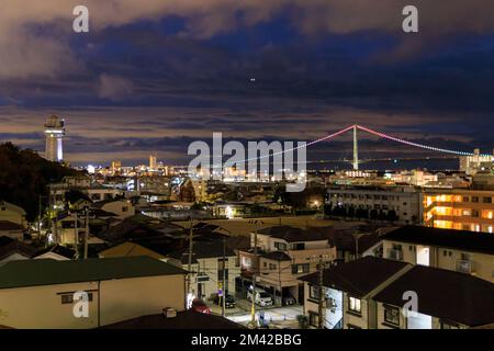 Luci colorate sul ponte sospeso con luci dalla piccola città in un'ora blu Foto Stock