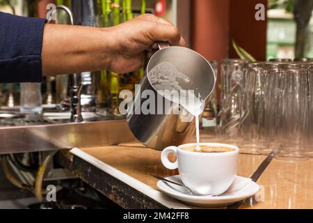 Alcune mani di un cameriere versano latte caldo in una tazza di caffè con una caraffa metallica in un bar Foto Stock