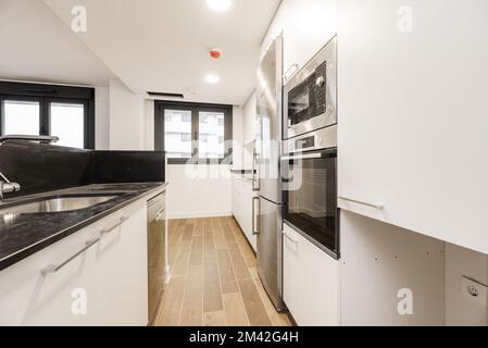 cucina con un'isola con un piano in marmo nero, una colonna di elettrodomestici in acciaio inossidabile, mobili in legno bianco e pavimenti in gres simile al legno Foto Stock