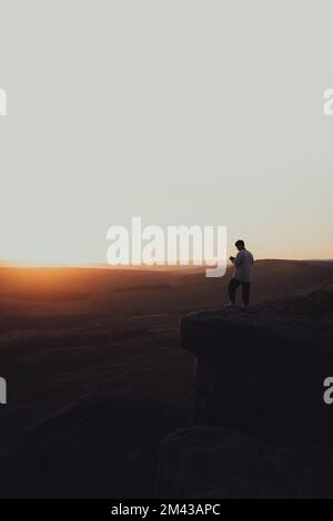 Un colpo verticale della persona che sta in piedi al bordo di una collina rocciosa ad un tramonto ipnotico Foto Stock