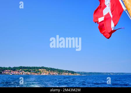 Bandiera danese a poppa di una barca da escursione e vista pittoresca della costa di Gudhjem, Bornholm isola, Danimarca. Foto Stock