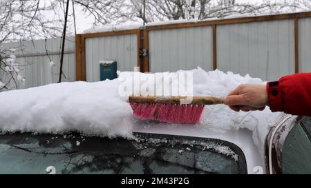 pulire uno spesso strato di neve dal tetto di un'auto grigia con una spazzola con un manico di legno in mano di un uomo Foto Stock