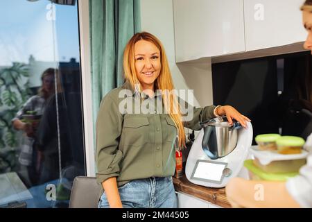 Donna bionda matura sorridente e sicura che tiene nuovo mixer o frullatore con recipiente in metallo in cucina accogliente. utensili da cucina moderni Foto Stock