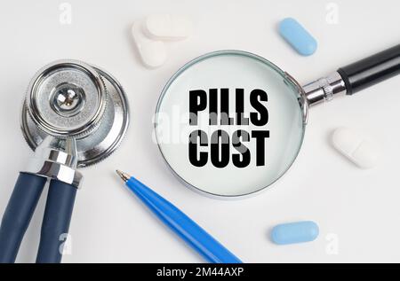 Medicina e concetto di salute. Su una superficie bianca si trovano pillole, una penna, uno stetoscopio e una lente d'ingrandimento con l'iscrizione - COSTO PILLOLE Foto Stock