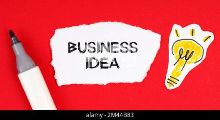 Concetto e idea di business. Su una superficie rossa si trovano un marcatore, un disegno di una lampadina e carta strappata con l'iscrizione - IDEA DI BUSINESS Foto Stock
