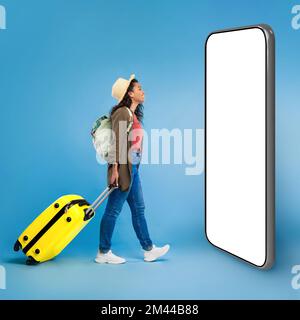 Felice giovane donna nera che cammina con valigia, controllando lo schermo dello smartphone Foto Stock