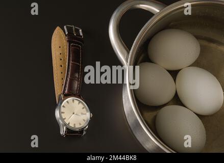 Tempo per lessare le uova. Uova di pollo in una padella e un orologio da polso su un pannello scuro Foto Stock