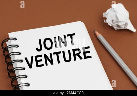 Concetto di business e finanza. Su una superficie marrone giace una penna, carta sgualcita e un blocco note con l'iscrizione - Joint Venture Foto Stock