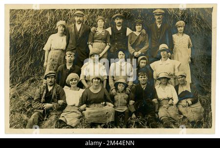 Originale e affascinante fine degli anni '20 primi anni '30 's cartolina di allegro ma stanco cercando lavoratori classe operaia che si rilassano per una fotografia di gruppo dopo il raccolto è stato portato in, grande pagliaio dietro di loro, un sacco di personaggi, giovani uomini e donne, alcuni bambini, membri della famiglia. I vestiti dei lavoratori sono protetti da grembiuli di tela di sacco. Fotografia di Ashby Swift, South Street, Bourne, South Lincolnshire, Inghilterra, REGNO UNITO Foto Stock