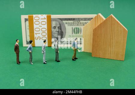 Concetto economico. Sulla superficie verde sono case di legno, una pila di dollari e figurine in miniatura di persone in piedi in linea per acquistare una casa. Foto Stock