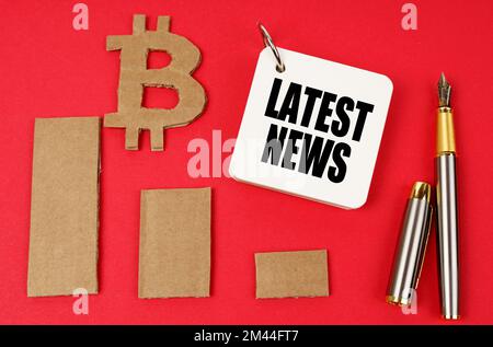 Business e bitcoin concetto. Su una superficie rossa si trovano un simbolo di bitcoin, un grafico, una penna e un blocco note con l'iscrizione - ULTIME NOTIZIE Foto Stock