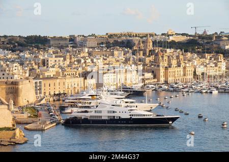 Birgu, Malta - 13 novembre 2022: Paesaggio urbano con facciate in pietra e forte che splende al tramonto e grande porto turistico con yacht ormeggiati e nave a vela Foto Stock