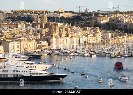 Birgu, Malta - 13 novembre 2022: Paesaggio urbano con facciate in pietra che brillano al tramonto e grande porto turistico con yacht ormeggiati e navi a vela Foto Stock