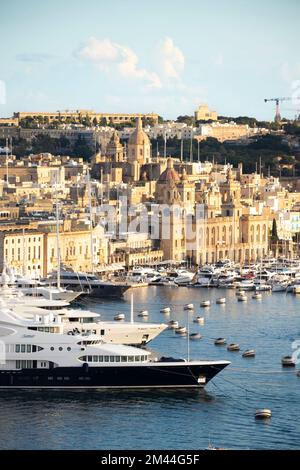 Birgu, Malta - 13 novembre 2022: Paesaggio urbano con facciate in pietra che brillano al tramonto e grande porto turistico con yacht ormeggiati e navi a vela Foto Stock