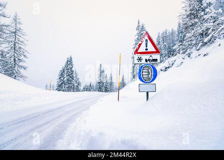 Cartello segnaletico e un cartello con catene di pneumatici lungo una strada innevata di montagna durante una tempesta di neve Foto Stock