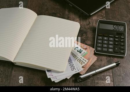 Frammento di libro contabile di un dispositivo di conto con banconote in euro e una penna stilografica sullo sfondo di un tavolo di legno. Foto Stock