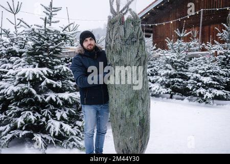Giovane uomo in piedi accanto all'albero di Natale. Albero grande per la famiglia grande. Buone vacanze invernali Foto Stock