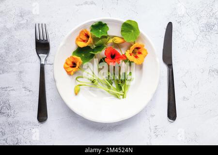 Piante di nasturzio, fiori e foglie di crescione indiane in piatto, posate su tavolo bianco, posate in piano Foto Stock