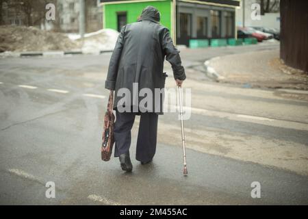 Vecchia donna cammina giù per strada in giacca nera. Pensionato in Russia sulla strada. Nonna con bastone da passeggio. Foto Stock