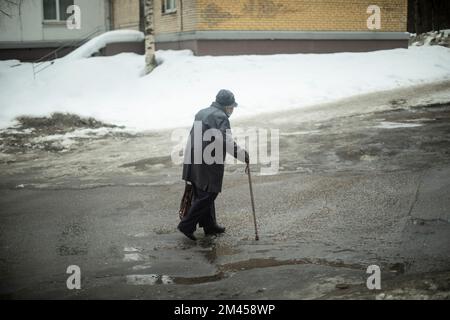 Vecchia donna cammina giù per strada in giacca nera. Pensionato in Russia sulla strada. Nonna con bastone da passeggio. Foto Stock