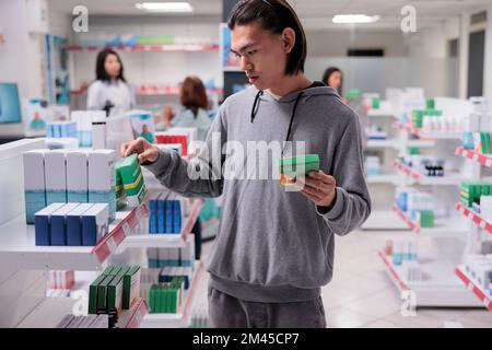 Cliente asiatico maschile esaminare supplementi sugli scaffali, cliente guardando le scatole di pillole in farmacia riempito con prodotti per la salute come vitamine e farmaci. Ricerca di forniture mediche. Foto Stock