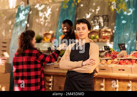 Allegro giovane agricoltore in posa accanto alla bancarella del mercato degli agricoltori, presentando prodotti alimentari naturali sani all'esterno. Donna fiduciosa che vende prodotti biologici di stagione, frutta e verdura. Foto Stock