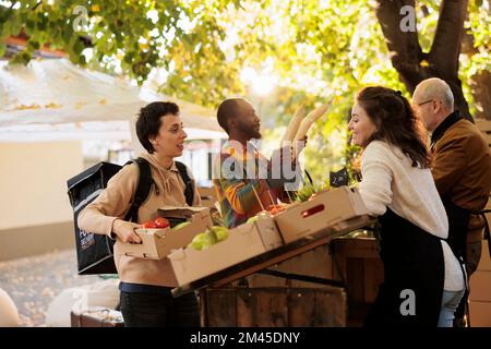 Allegro corriere e proprietario locale stand parlare di prodotti naturali, wokring sulla consegna di cibo fresco ordine ai clienti. Greengrocer dando scatola con frutta e verdure alla donna di consegna. Foto Stock