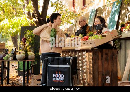 Giovane donna con borsa termica che raccoglie l'ordine al mercato locale, consegnando cibo naturale. Corriere femminile in piedi vicino allo stand dei prodotti agricoli, che prende frutta e verdura biologica fresca. Foto Stock