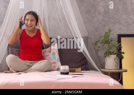 Allegra ragazza adolescente che ascolta musica sulle cuffie mentre si siede a letto Foto Stock