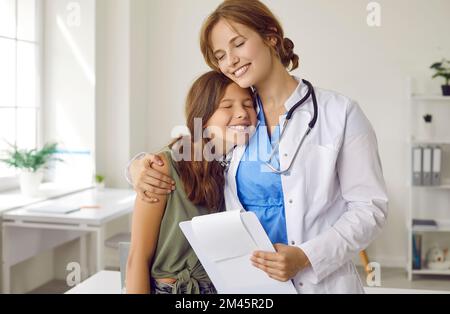 Felice pediatra in piedi in ufficio medico, abbracciando paziente bambino e sorridendo Foto Stock