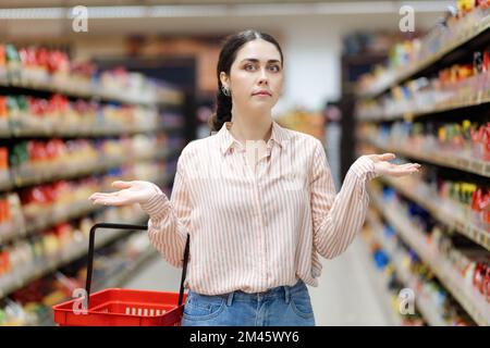 Ritratto di stupore giovane donna caucasica tiene il carrello della spesa e le distende le braccia fuori ai lati. Scaffali del negozio di alimentari e corridoio sullo sfondo. Conc Foto Stock