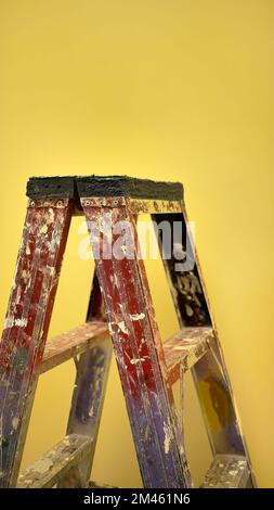 Vecchia scala di pittura usata costruzione con spruzzi dipinti che si erette di fronte alla parete gialla sotto calda luce bianca, verticale Foto Stock