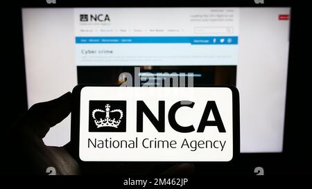 Persona che tiene il cellulare con il logo della British National Crime Agency (NCA) sullo schermo di fronte alla pagina web. Messa a fuoco sul display del telefono. Foto Stock