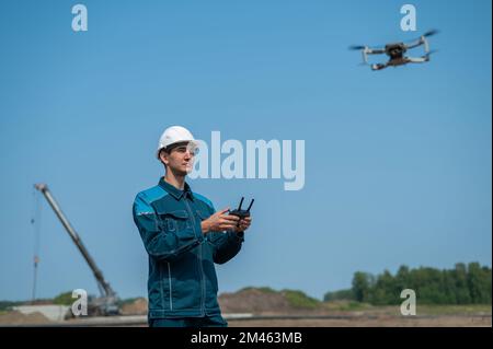 Un uomo in un casco e tute controlla un drone in un cantiere. Il costruttore esegue la supervisione tecnica. Foto Stock