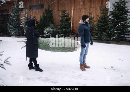 Figlio con la sua mamma portare l'albero di Natale alla macchina. Conifere dal vivo da eco negozio in villaggio. Anticipazione della vacanza. Umore invernale Foto Stock