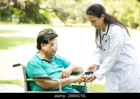 Controllo medico indiano con pulsossimetro dispositivo sul dito di un paziente anziano di sesso maschile diabetico in una sedia a rotelle all'aperto al parco. Foto Stock