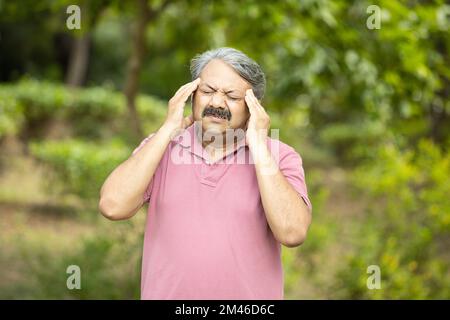 Uomo anziano indiano che soffre di mal di testa in piedi all'aperto al parco. Anziani stressati, Foto Stock