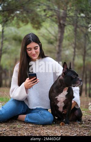 Giovane donna caucasica che usa il telefono seduto in una foresta con il suo cane . Foto Stock