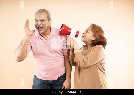 Donna indiana anziana urla o urla contro l'uomo in megafone isolato su sfondo beige. annunciare la vendita di sconti. Moglie asiatica tease marito. Foto Stock