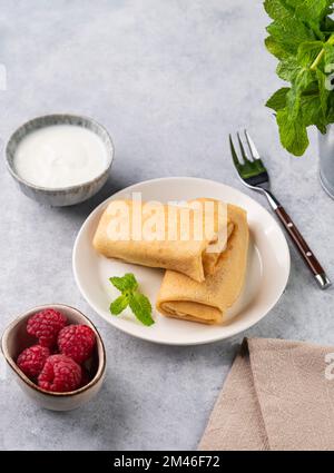 Crepe fatte in casa con lampone e panna acida su un piatto su uno sfondo chiaro con menta fresca. Concetto di cibo sano. Spazio di copia. Foto Stock