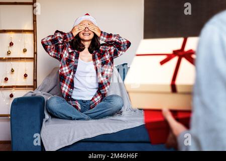 Giovane donna che indossa il cappello di Santa seduta sul divano con gli occhi chiusi con le mani mentre il suo ragazzo che porta le scatole del regalo di Natale. Foto Stock