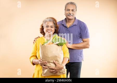Felice coppia indiana senior tenere sacchetti di carta della drogheria con cibo isolato su sfondo beige. foto da studio. Vita di pensionamento. Foto Stock