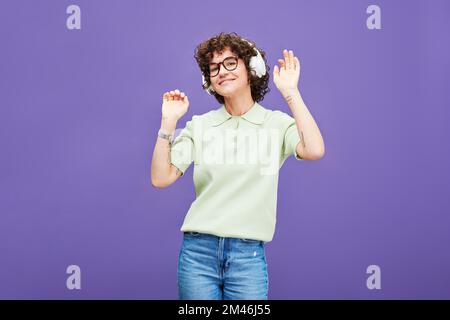 Giovane donna gioiosa in cuffie e casualwear ascoltando musica e ballando davanti alla macchina fotografica su sfondo lavanda in isolamento Foto Stock