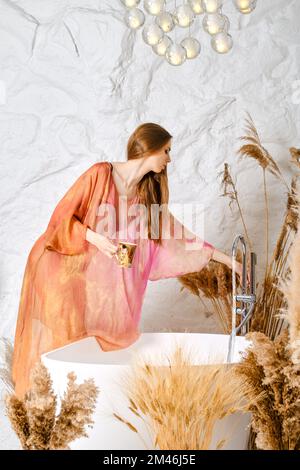 Donna in stola di seta riempie una vasca da bagno di acqua per rilassarsi Foto Stock