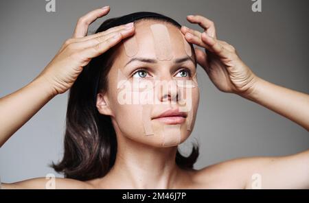 Giovane donna adulta che applica nastro di cinesiologia facciale sul suo viso, trattamento facciale della pelle con nastro adesivo. Foto Stock